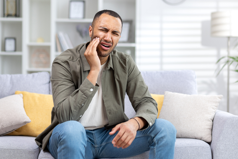 Young hispanic man suffering from toothache sitting on sofa at home and holding hand to cheek.